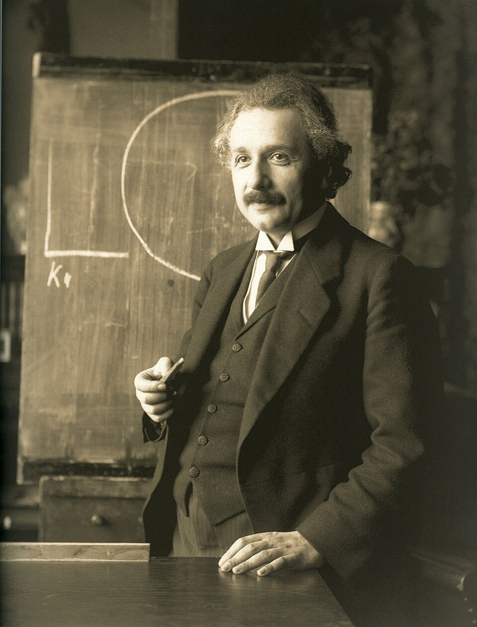 The 1921 sepia portrait of Albert Einstein in a three-piece suit, holding a piece of chalk beside a blackboard, one hand resting on a desk.