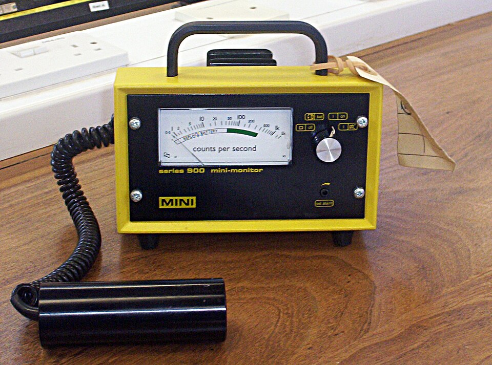 A portable Geiger-Muller counter: a yellow-and-black instrument box with an analogue dial marked 'counts per second' and a cylindrical detector probe on a coiled cable lying beside it.