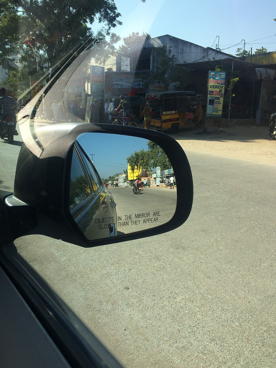 A car's outer rear-view mirror photographed from inside the car on an Indian street, its curved glass showing a wide shrunken view of the road behind with the words 'objects in the mirror are closer than they appear' etched across it.
