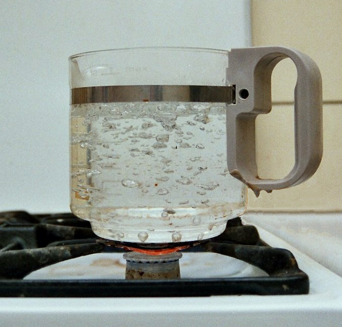 A glass measuring jug of water boiling over a gas flame, with clusters of vapour bubbles forming inside the water and rising to the surface.