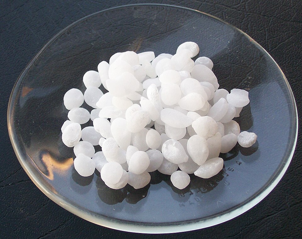 A heap of white, semi-translucent sodium hydroxide pellets resting on a round glass watch glass.