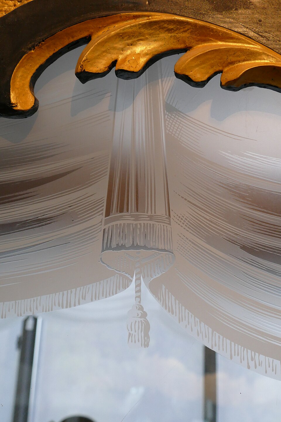 A close-up of a decorative etched glass pane in a gilt frame, the etched areas frosted matte white in a swagged curtain-and-tassel pattern against the clear glass behind.