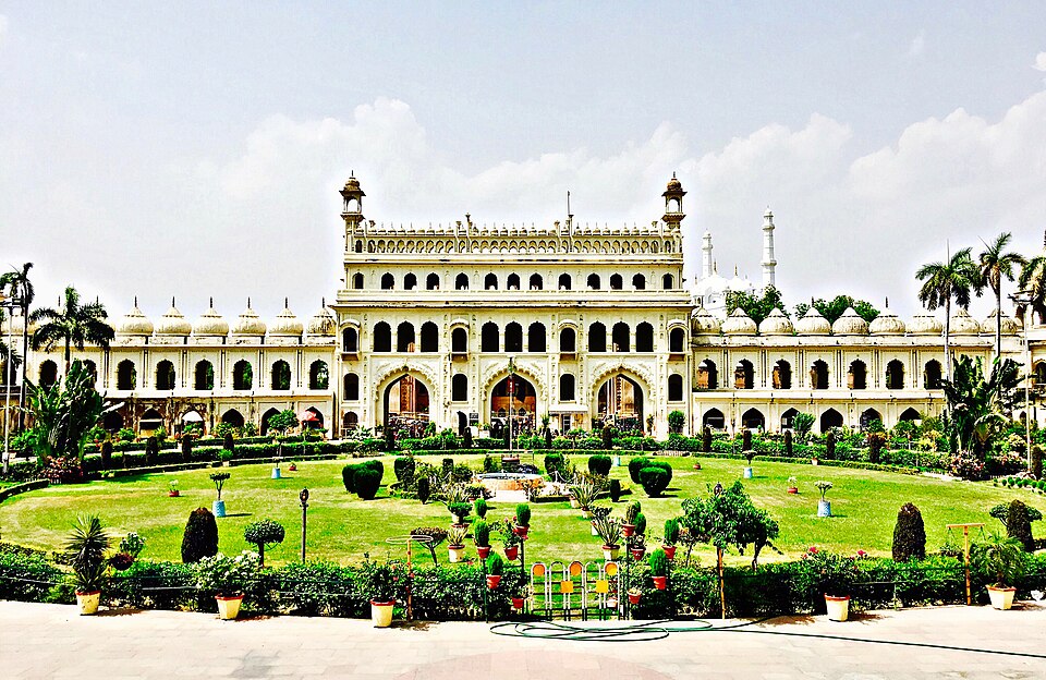The ornate cream-coloured gateway of the Bara Imambara complex at Lucknow seen across a formal lawn, with the white domes and minarets of the Asafi Mosque rising behind it.