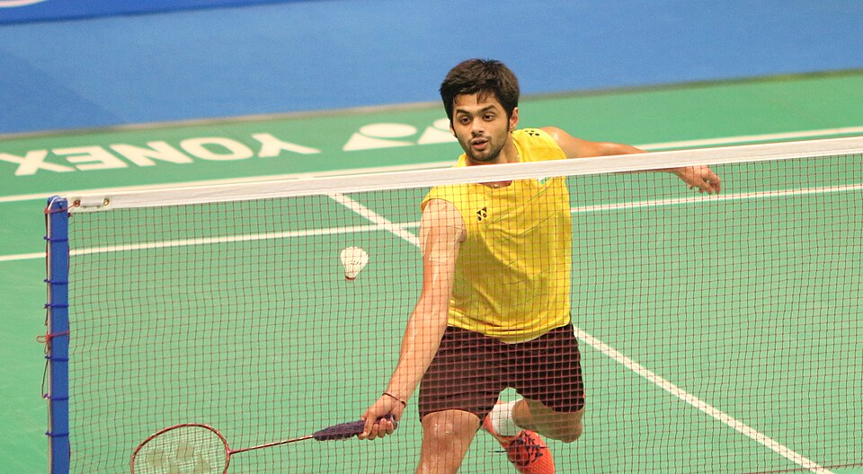 Indian badminton player B. Sai Praneeth in a yellow jersey playing a shot at the net during a match on a green indoor court.
