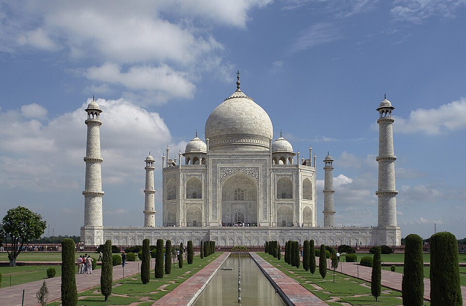 The white marble Taj Mahal at Agra with its central dome and four minarets, seen straight down the reflecting pool lined with clipped cypresses, with visitors on the terrace.