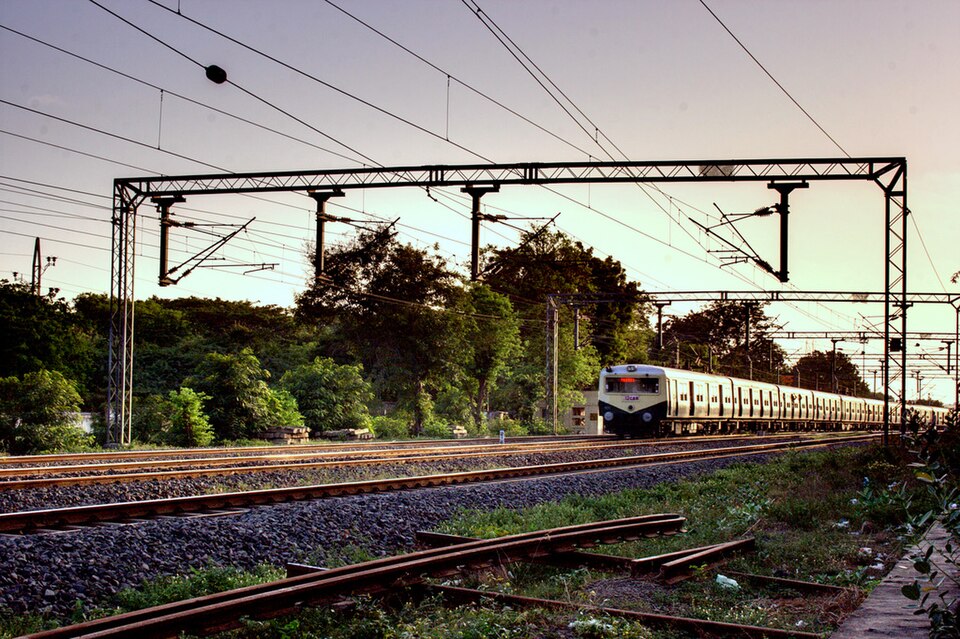 An electrified Indian Railways line at dusk, with overhead traction wires strung from a steel portal gantry above the tracks and a suburban EMU train approaching.