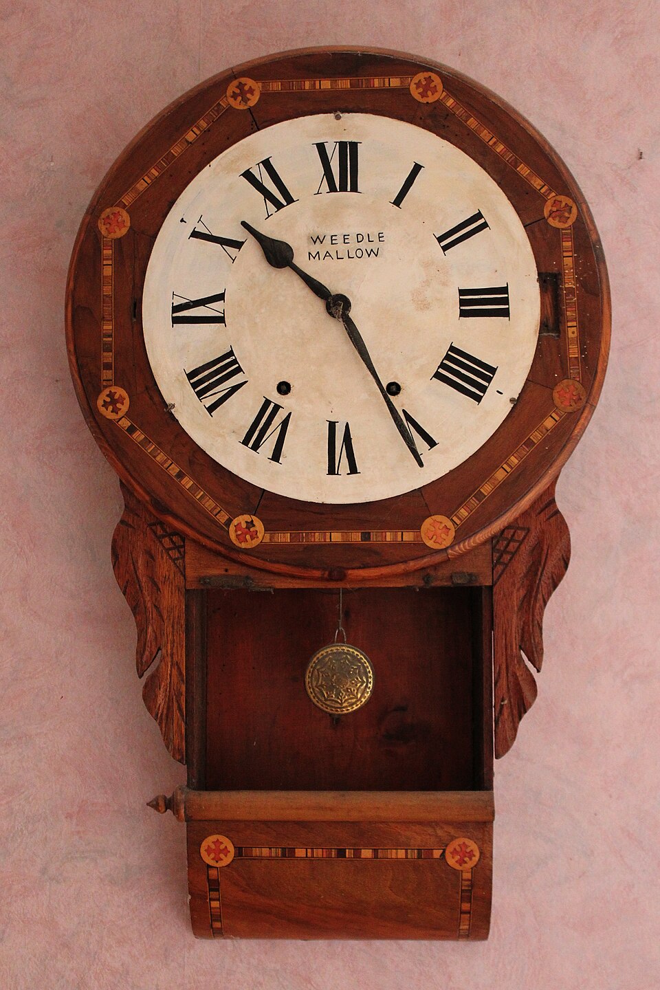 A carved wooden wall clock with a Roman-numeral dial and its brass pendulum bob hanging in the open case below.