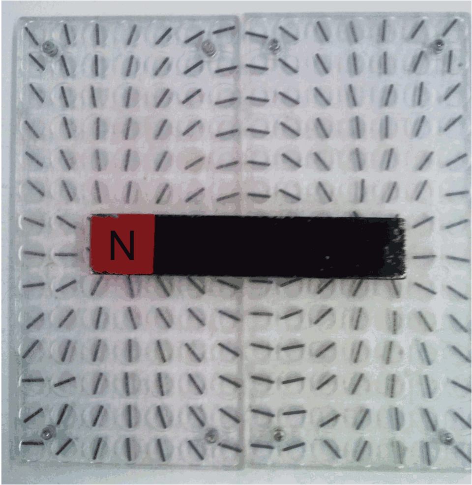 A bar magnet with its north pole marked in red lying on a board of small compass needles, which line up along the magnet's field.