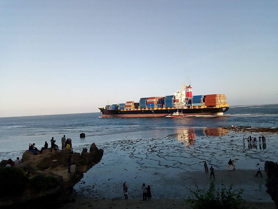A steel container ship stacked with cargo floats on the sea beside a small tug, seen from a rocky shoreline.