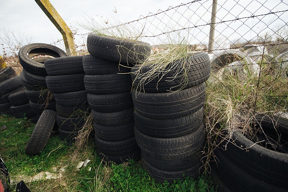 Stacks of worn black rubber car tyres piled up beside a wire mesh fence with grass growing between them.