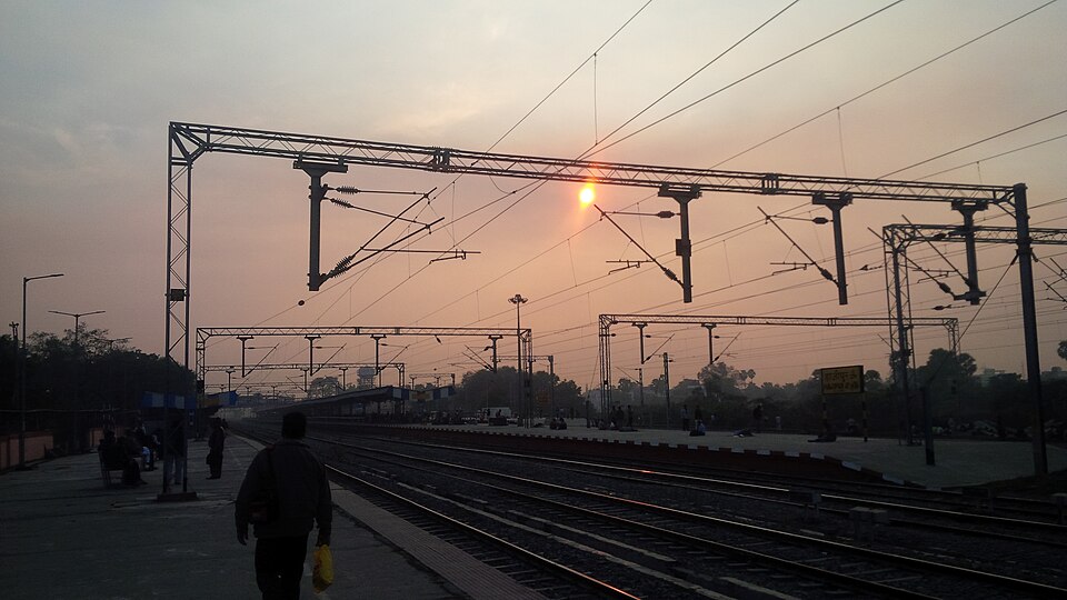 Hajipur Junction station in Bihar at sunset, with overhead electrification gantries spanning the tracks and passengers waiting along the platforms.