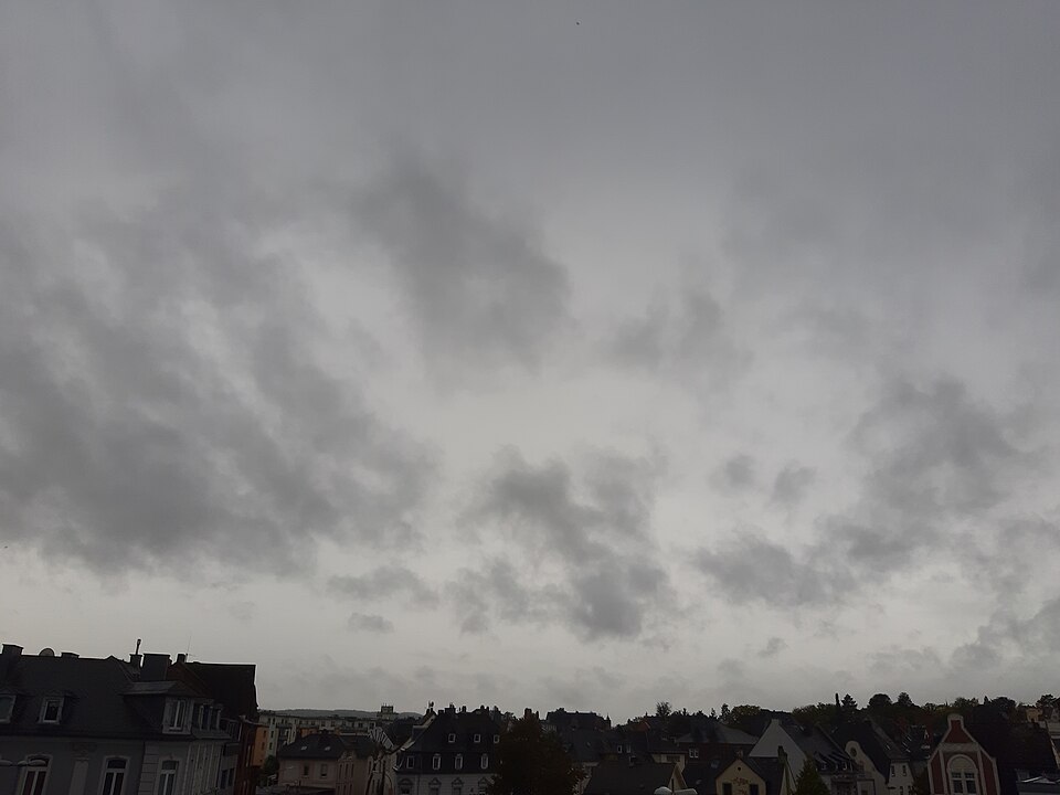 A thick, featureless dark grey cloud layer covering the whole sky above a town’s rooftops, with ragged lower shreds of cloud drifting beneath it.