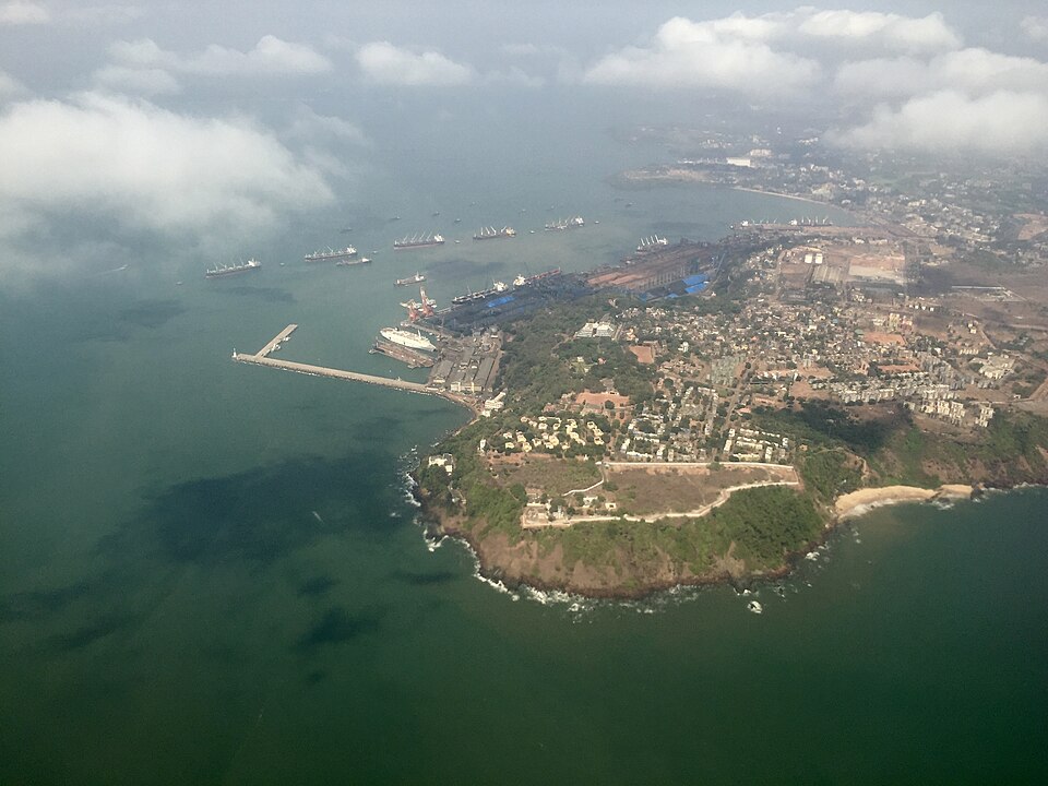 Aerial view of Mormugao harbour on a green headland, with a long breakwater arm, port berths and cranes, and cargo ships lying at anchor offshore.