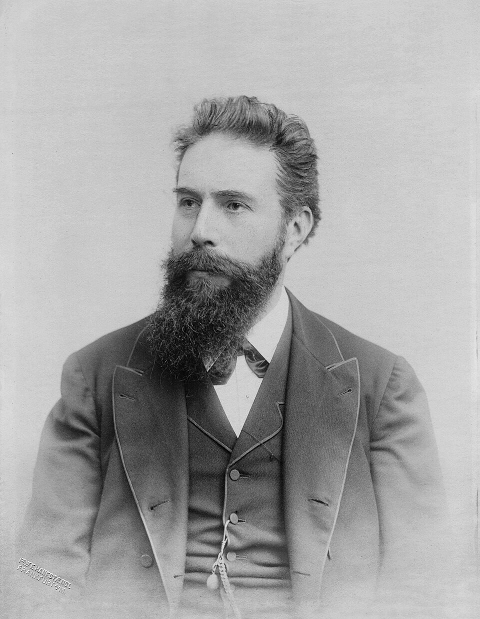 Black-and-white studio portrait of Wilhelm Conrad Röntgen as a middle-aged man with a full dark beard and swept-back hair, wearing a three-piece suit and bow tie.