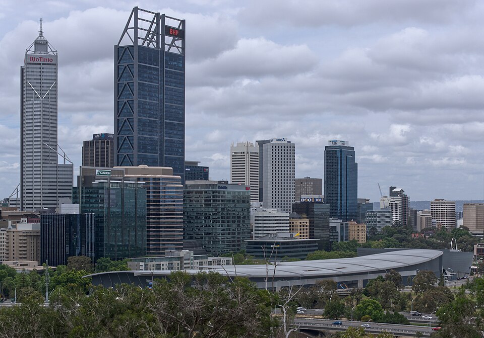 Daytime view over the Perth central business district in Western Australia, with a cluster of glass and concrete office towers rising above green parkland under a cloudy sky.