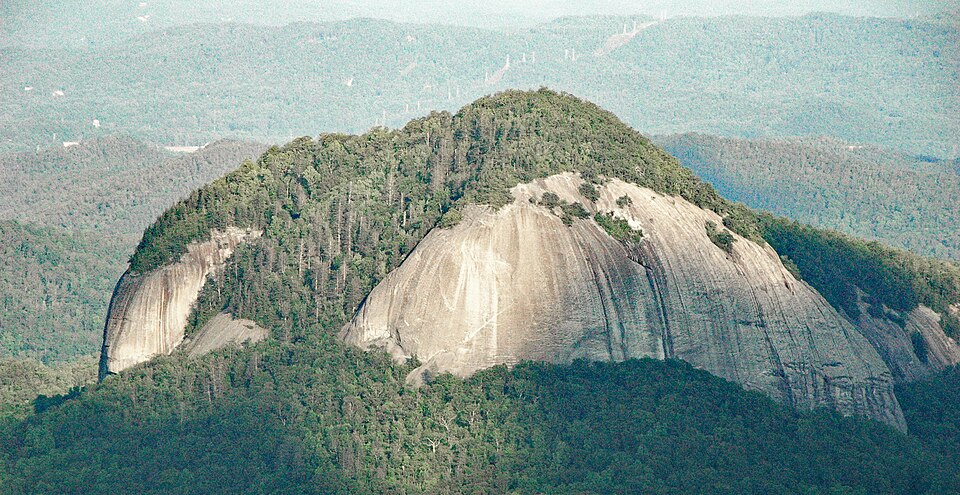 A bare rounded dome of granite rising out of forested hills, its pale face shedding curved slabs of rock in long vertical sheets.
