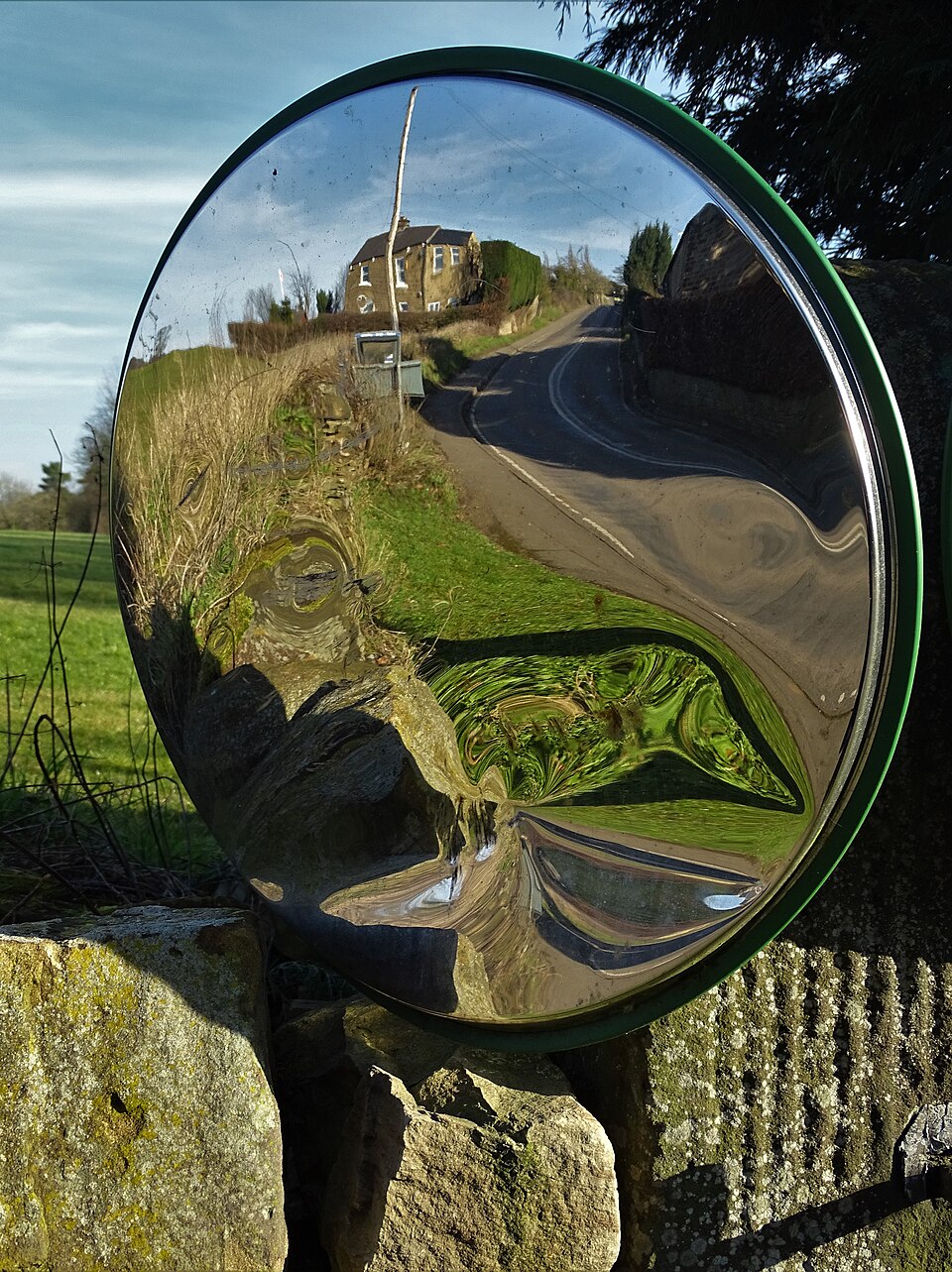 A round convex road-safety mirror fixed on a stone wall beside a country lane, its bulging surface showing an upright, wide-angle view of the road, verge and a house beyond the bend.