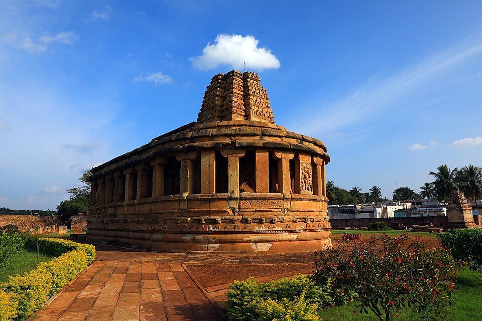 The Durga temple at Aihole in Karnataka, a sandstone shrine on an apsidal plan ringed by a pillared verandah and topped by a stepped tower, standing on a lawn under a blue sky.