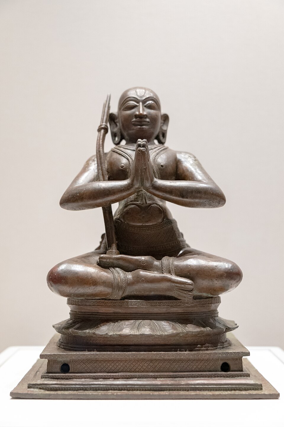 A South Indian bronze figure of the teacher Ramanuja, seated cross-legged on a lotus base with his palms joined in reverence and a slender three-staffed tridandi resting against his shoulder.