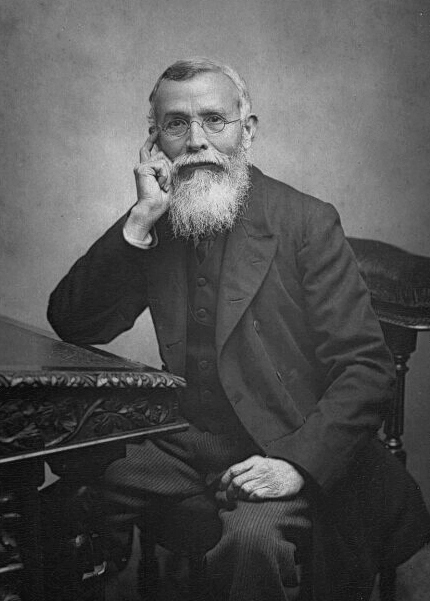 Black-and-white studio photograph of an elderly bespectacled man with a full white beard in a dark suit, seated beside a carved table with his chin resting on one hand.