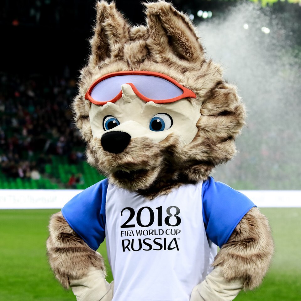 A costumed grey wolf mascot with red sports goggles and a blue-and-white shirt reading '2018 FIFA World Cup Russia', standing hands on hips on a football pitch.