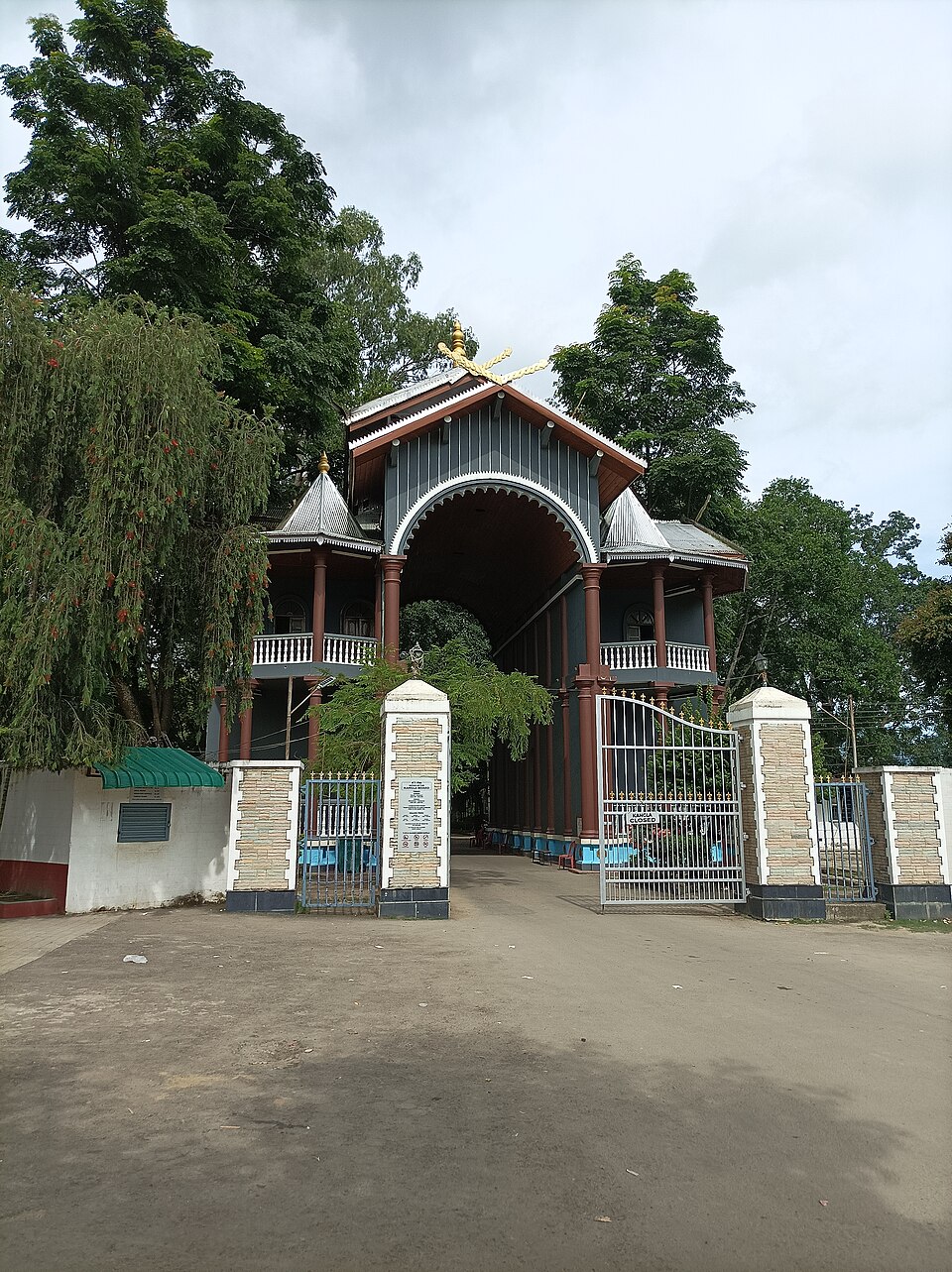 The Kangla Grand Gate in Imphal, a dark timber gatehouse with a wide arch, red pillars, twin conical turrets and a gilded finial, seen past open white iron gates at the end of a bare forecourt.
