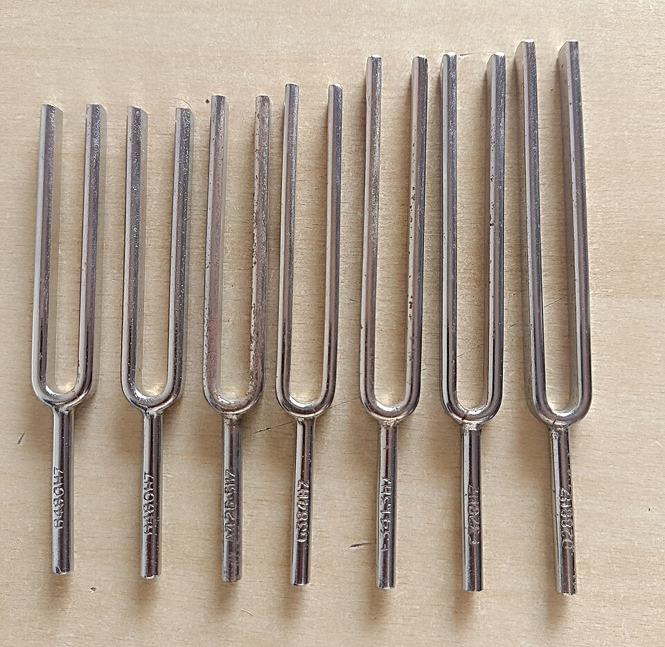 Seven steel tuning forks of different lengths laid side by side on a wooden surface, each handle stamped with its frequency in hertz.