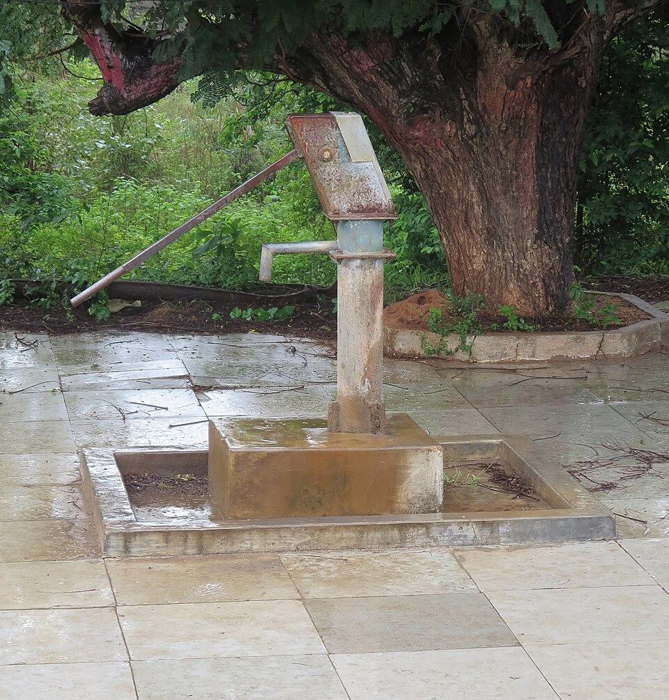 A metal hand pump standing on a concrete plinth with a drainage apron, beside a large tree, used for drawing groundwater.