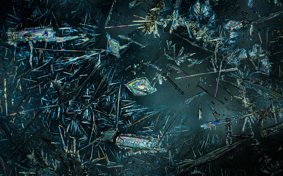 Sodium carbonate crystals photographed under a polarising microscope, showing iridescent needle- and plate-shaped crystals on a dark field.