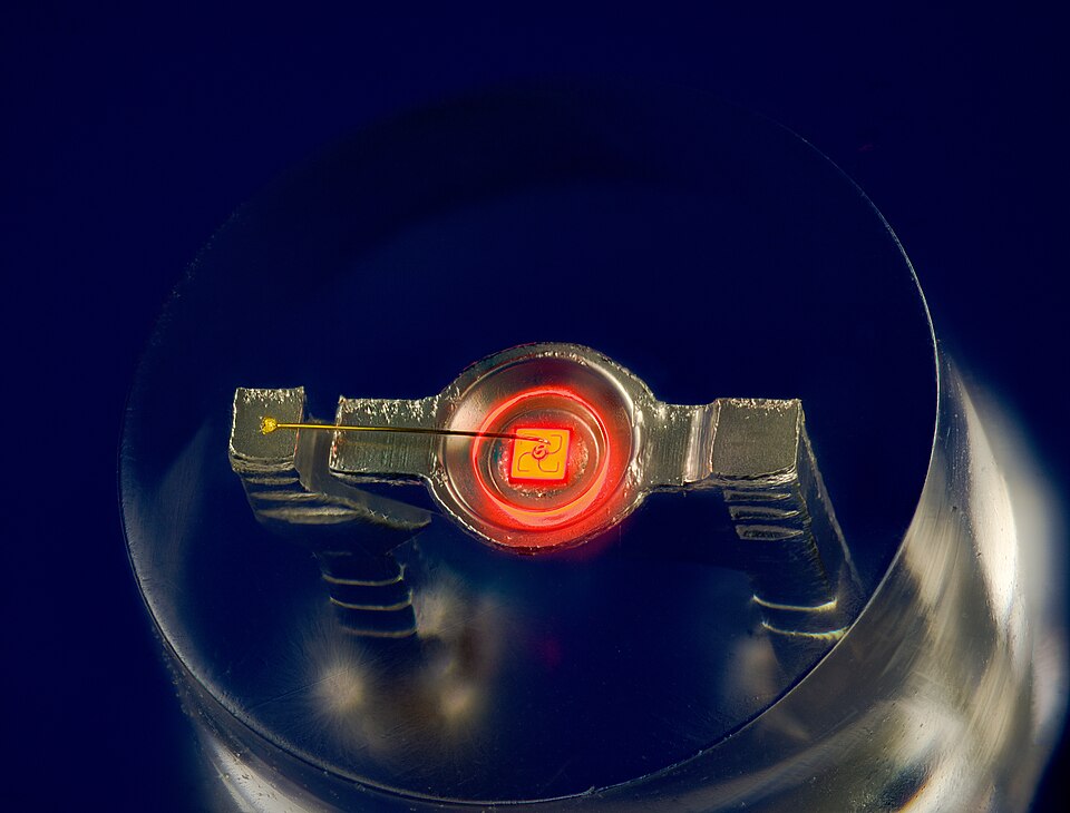 Close-up inside a lit red LED, showing the tiny square semiconductor chip glowing red in its metal reflector cup with a fine bond wire running to the other lead.