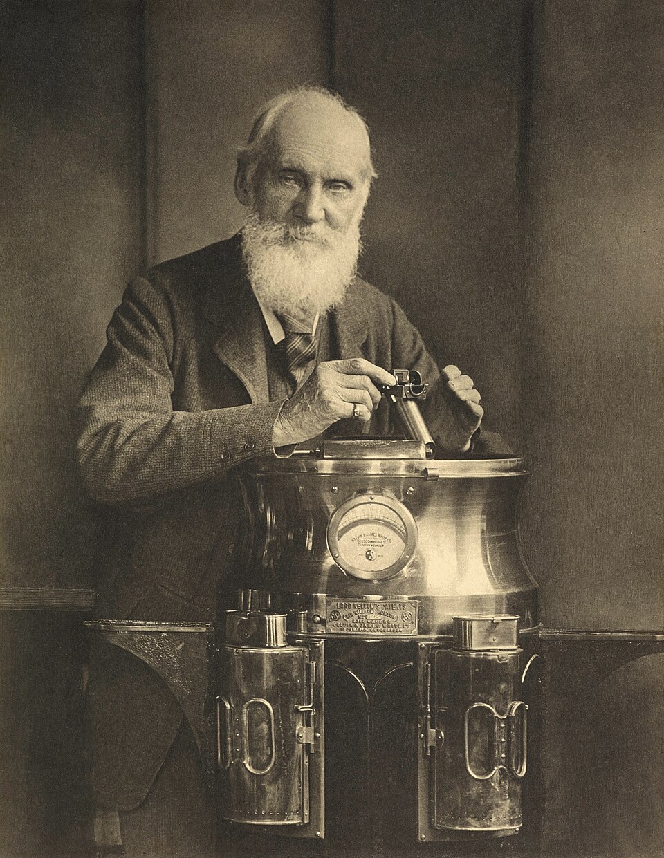 Sepia studio portrait of the elderly, white-bearded William Thomson, Lord Kelvin, standing behind a brass mariner's compass binnacle bearing his patent plate.