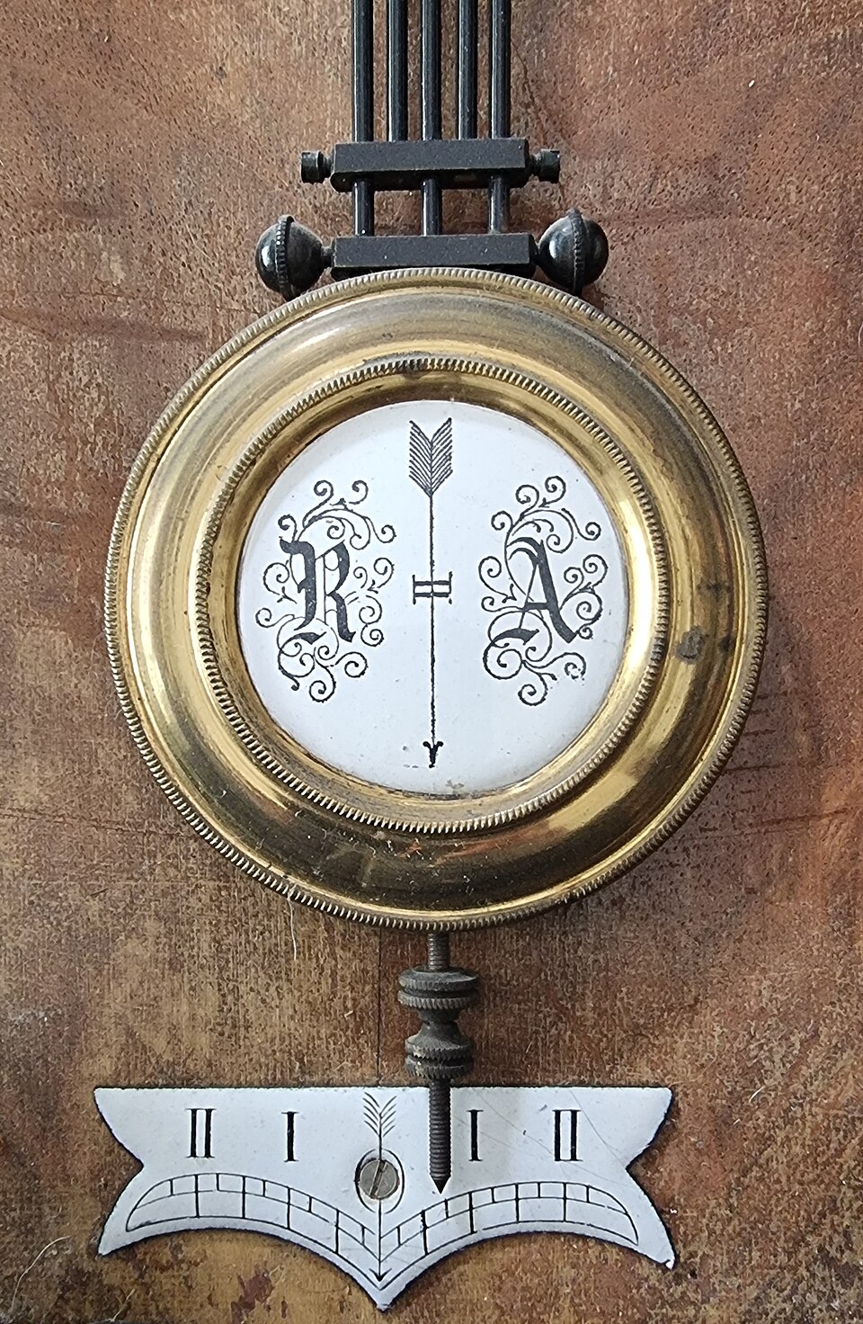 The heavy brass lens-shaped bob of a clock pendulum hanging on its rod, with the knurled rating nut below it and a small beat scale behind.