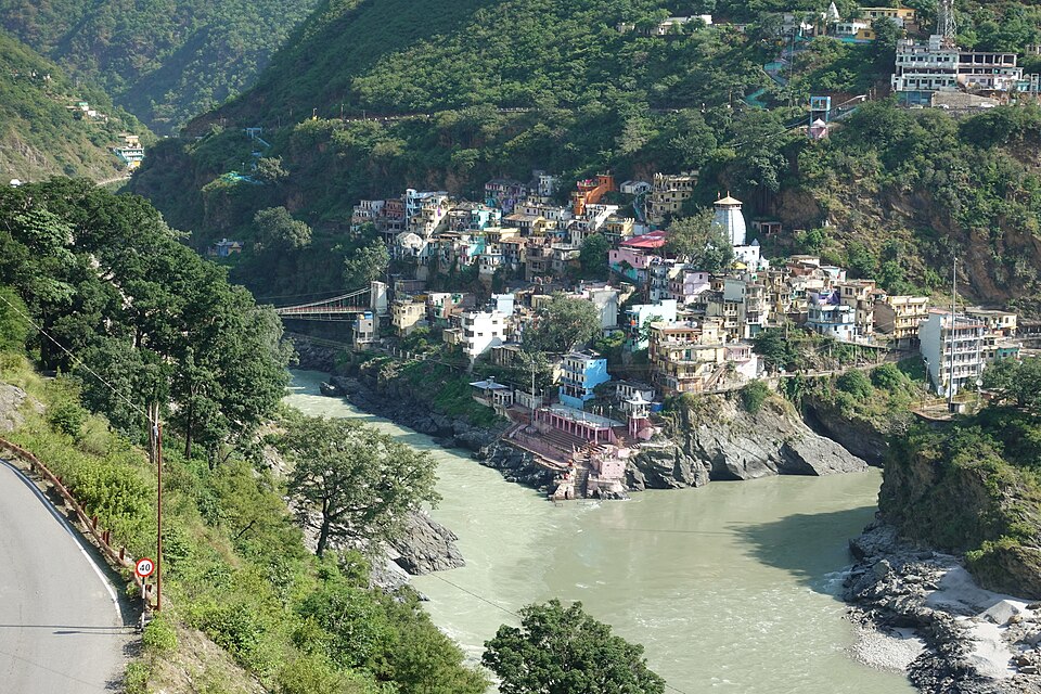 Aerial view of Devprayag town on a steep Himalayan spur, with two river channels meeting below it at a rocky point, ghats at the water’s edge and a suspension footbridge across one arm.