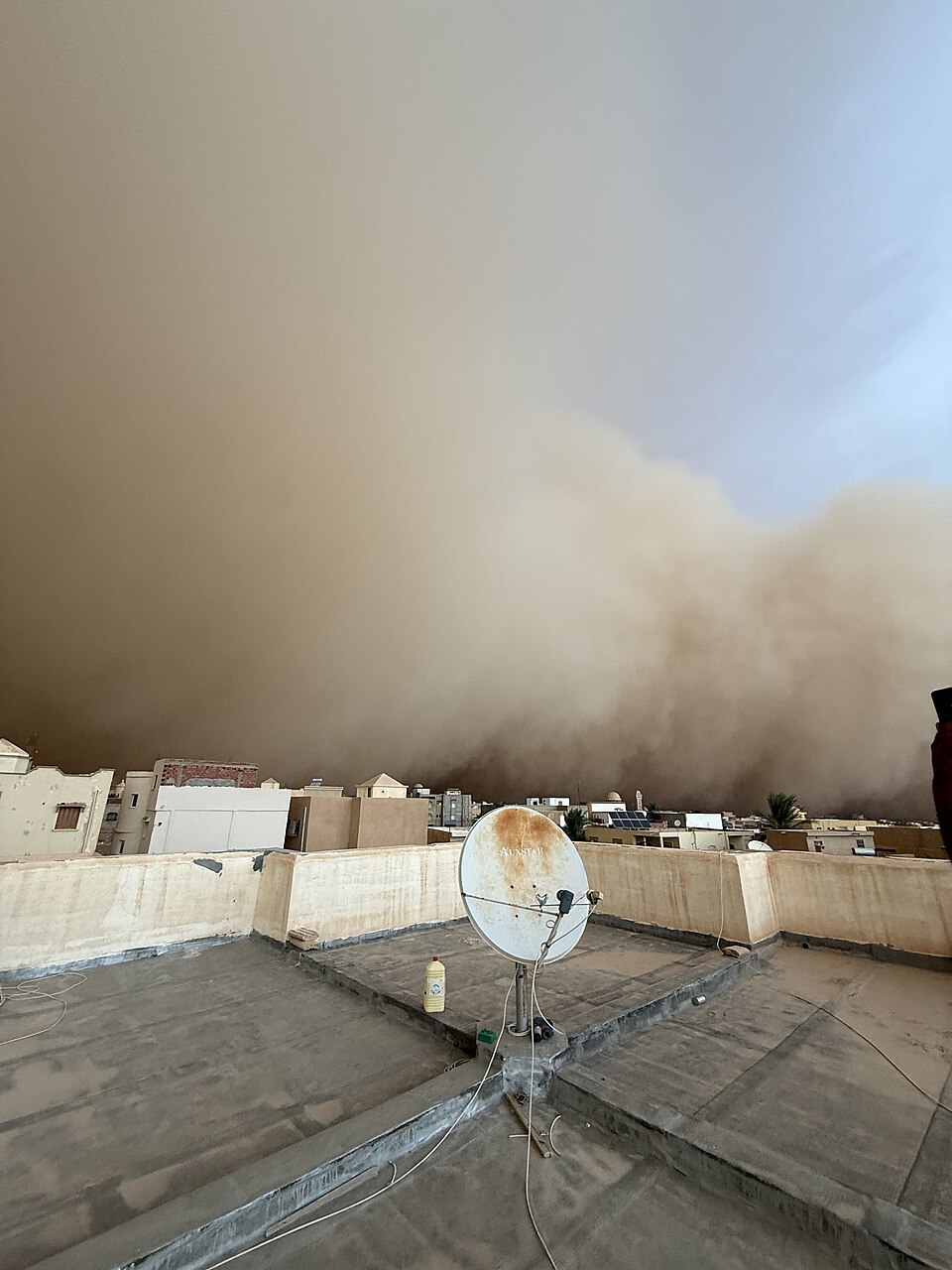 A towering tan wall of dust rolls in over a low desert town, seen from a flat rooftop, blotting out the sky above the houses.