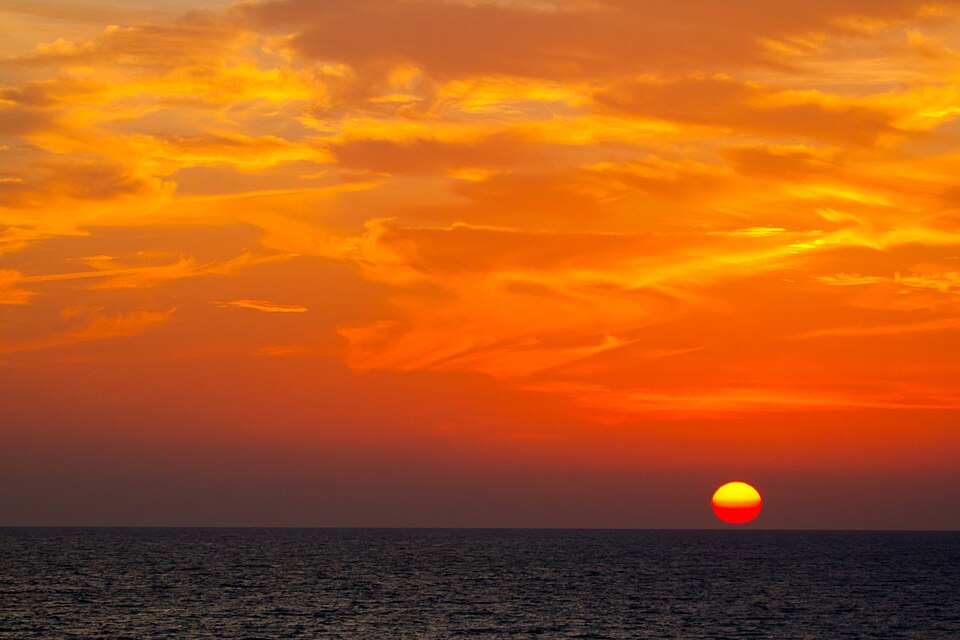 A deep red Sun sitting low on the sea horizon at sunset amid an orange sky