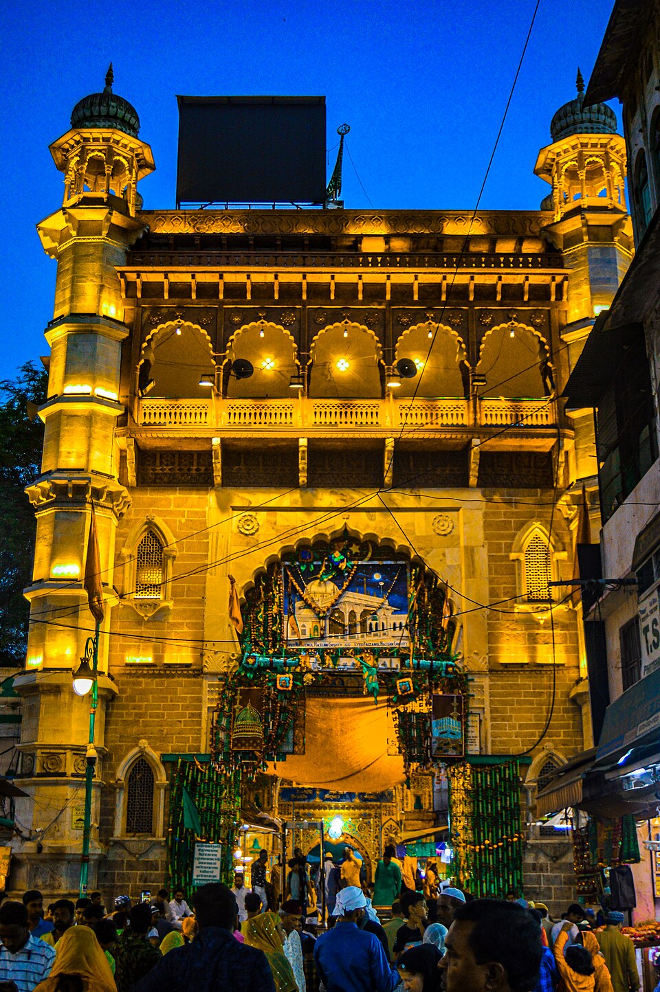 Illuminated ornate gateway of the Ajmer Sharif Dargah, the Chishti sufi tomb-shrine, with pilgrims below at dusk
