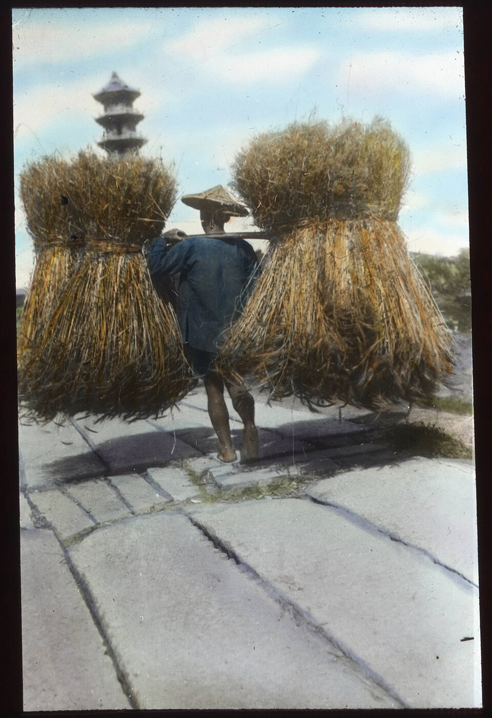 A labourer carrying two heavy bundles of straw on a shoulder pole, applying force to move a load.