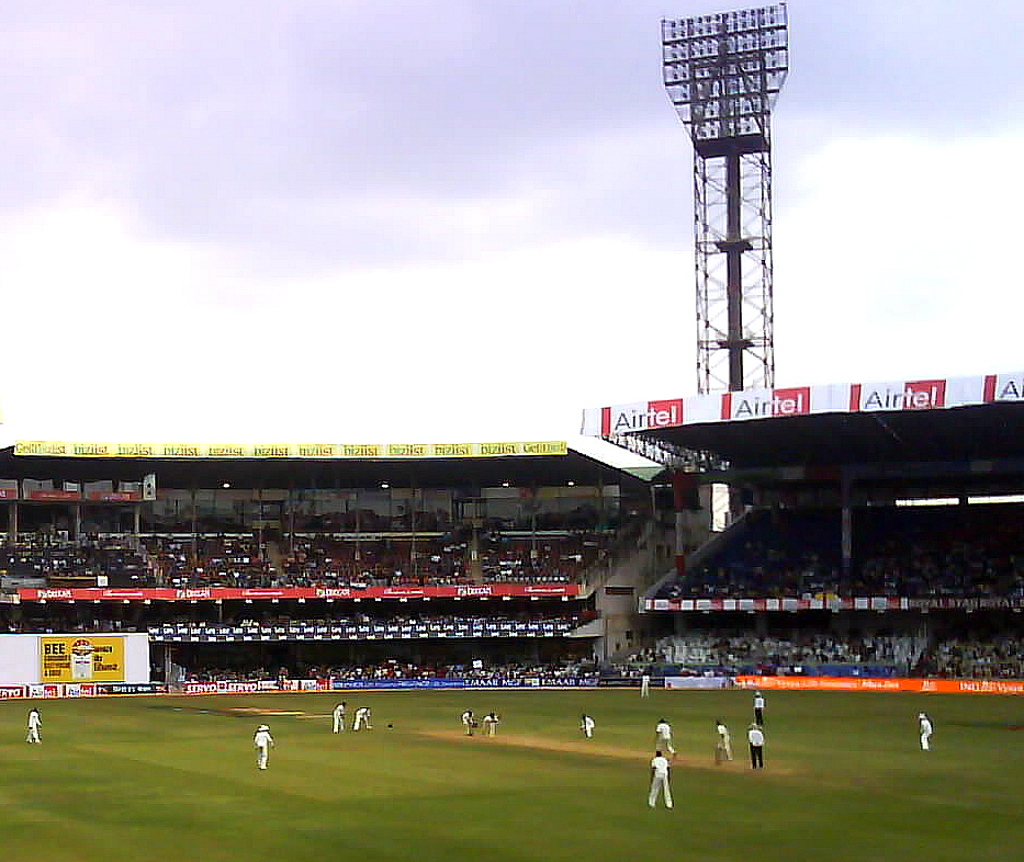 A Test cricket match in progress at a floodlit stadium, players in whites on the field