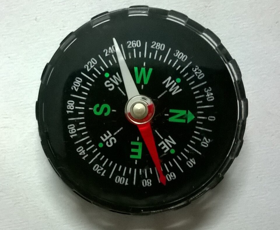 A small school-laboratory magnetic compass seen from above, its red-and-white magnetic needle balanced on a central pivot over a dial marked with the cardinal directions and degrees.