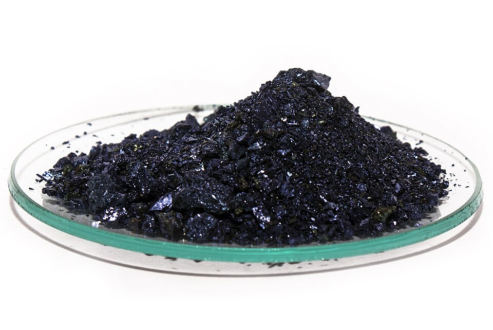 A heap of dark purple, almost black glinting potassium permanganate crystals on a glass watch glass against a white background.
