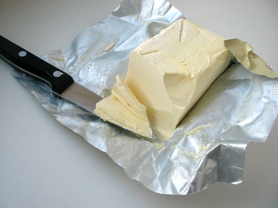 A block of pale-yellow solid margarine on its opened foil wrapper, with a knife cutting a slice from one end.