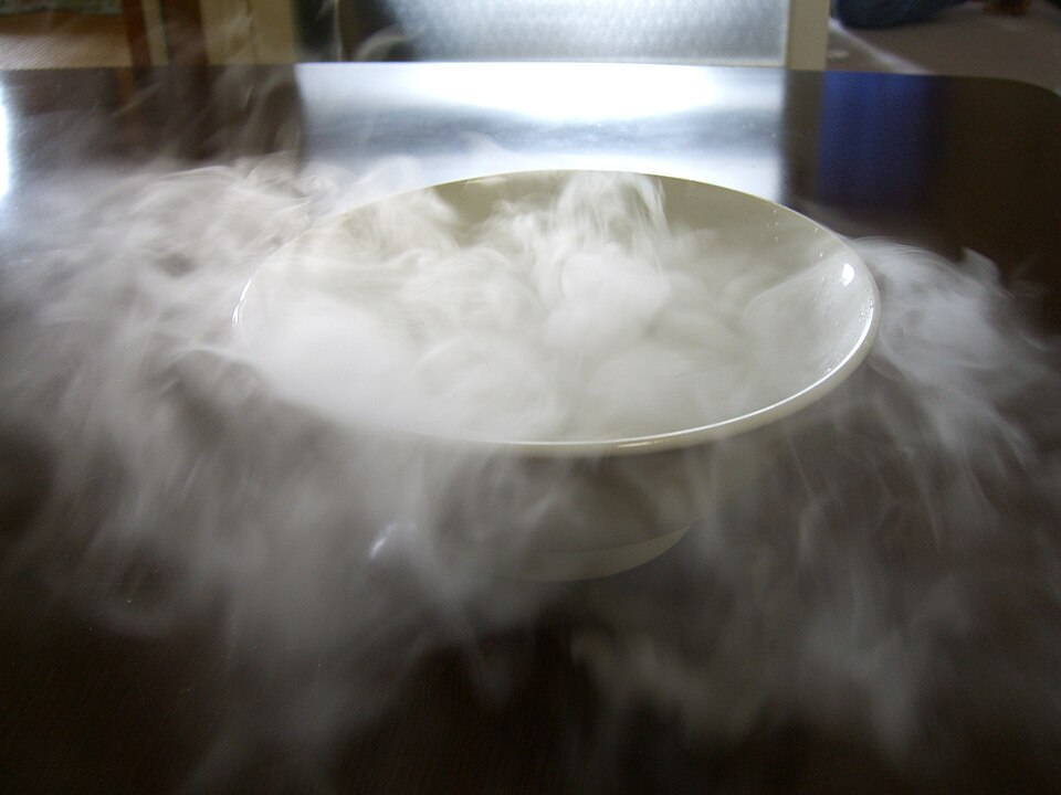 A white bowl of dry ice on a table, with thick white fog boiling out of it and spilling over the tabletop.