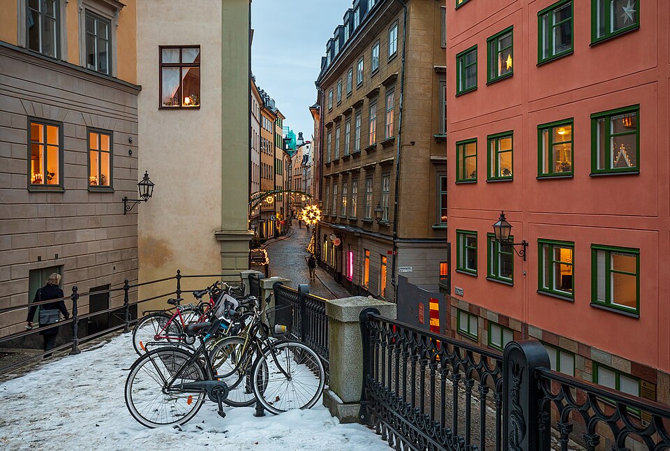 A narrow cobbled lane between tall ochre and red old town houses in Gamla Stan, Stockholm, with snow on the ground and bicycles parked by an iron railing.