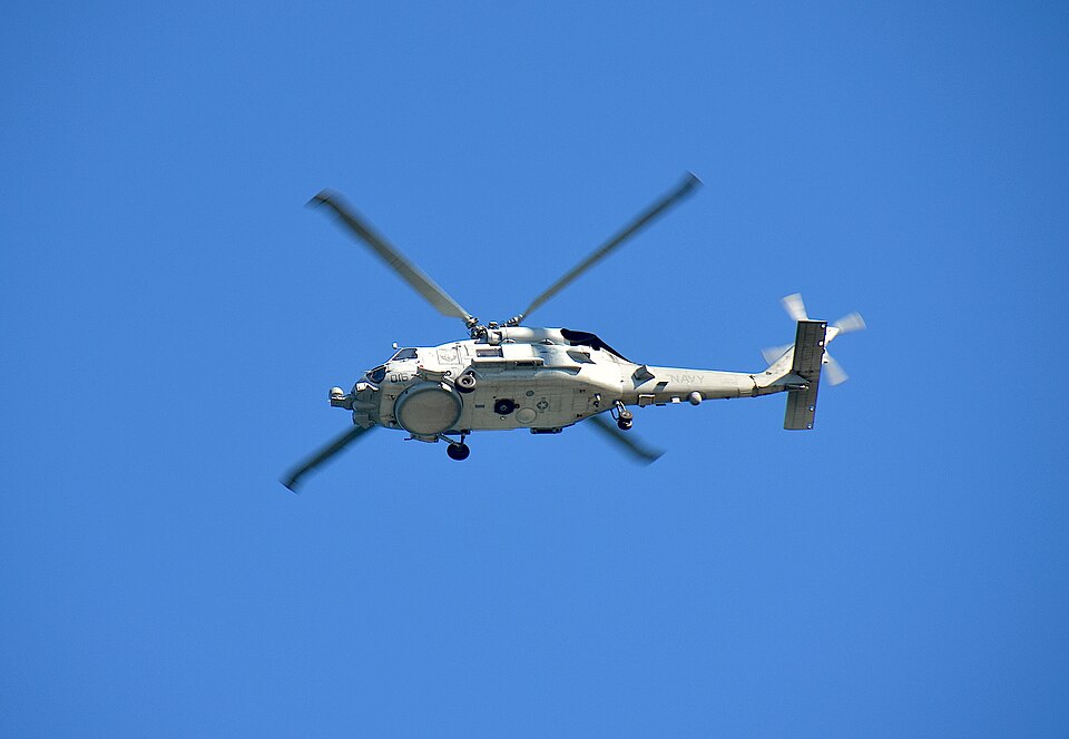 A grey MH-60R Seahawk naval helicopter marked NAVY flying against a clear blue sky, seen from below in side profile.