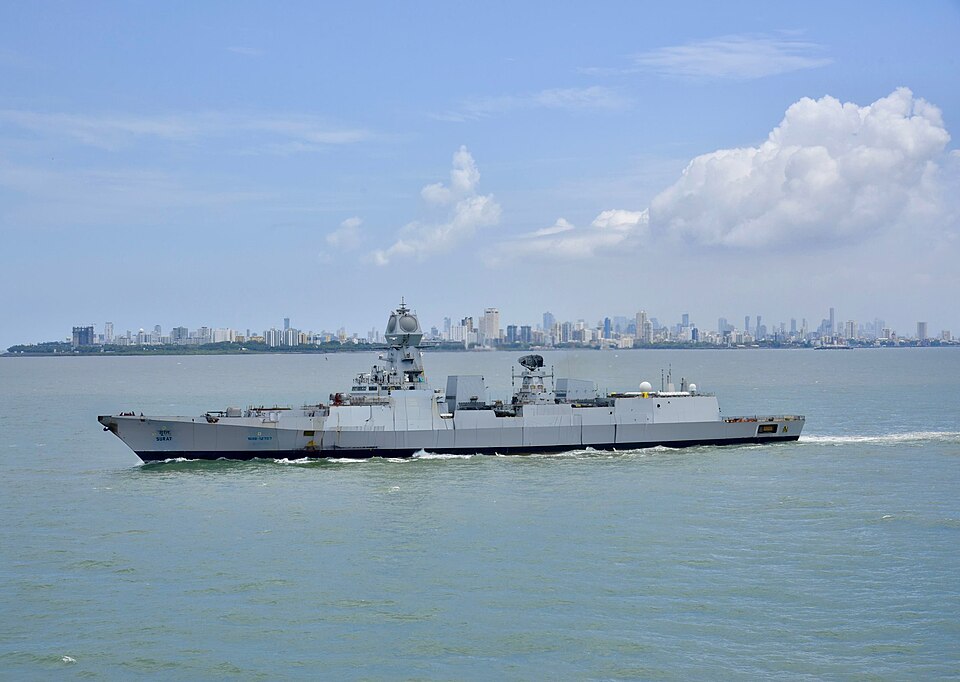 The Indian Navy stealth destroyer INS Surat under way at sea, its name painted on the bow, with the Mumbai skyline on the horizon behind.