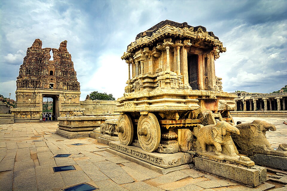 The carved stone chariot in the Vittala temple complex at Hampi, standing on its stone wheels with elephant sculptures in front and a ruined gateway tower behind.