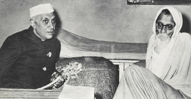 A black-and-white photograph of two men seated on a low platform: Jawaharlal Nehru on the left in a white Gandhi cap and dark coat, and Vinoba Bhave on the right, white-bearded and in round spectacles with a white cloth over his head, at Bhave's Paunar Ashram.