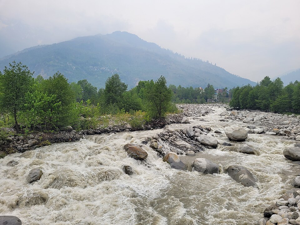 The Beas River running fast and white over boulders near Manali, with wooded Himachal hills rising behind it.