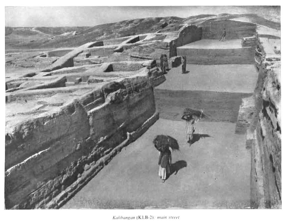 Archival black-and-white excavation photograph of the main street at the Harappan site of Kalibangan, running between exposed brick house walls, with workers carrying baskets of earth along it.