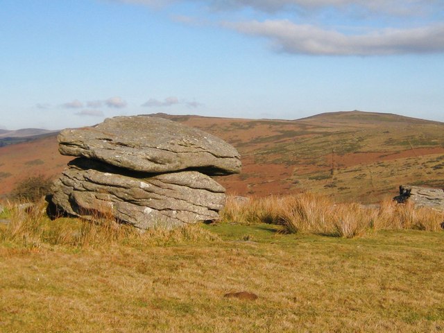 A free-standing granite tor on Dartmoor — a stack of horizontally jointed weathered granite blocks sitting on open moorland grass, with rolling moor behind.